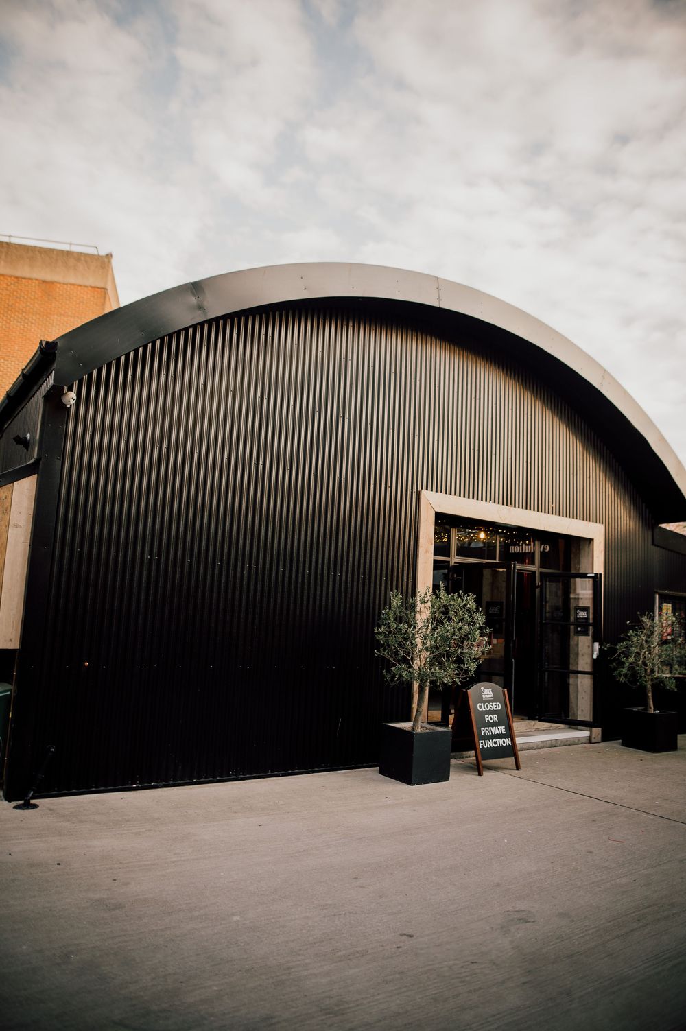 Modern black curved building exterior with arched entrances and concrete walkway under cloudy sky.