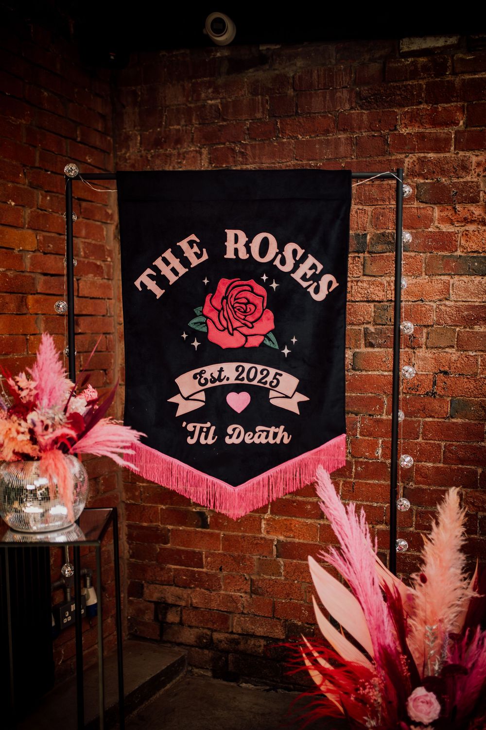 Pink neon sign reading 'The Roses' with rose design mounted on exposed brick wall.