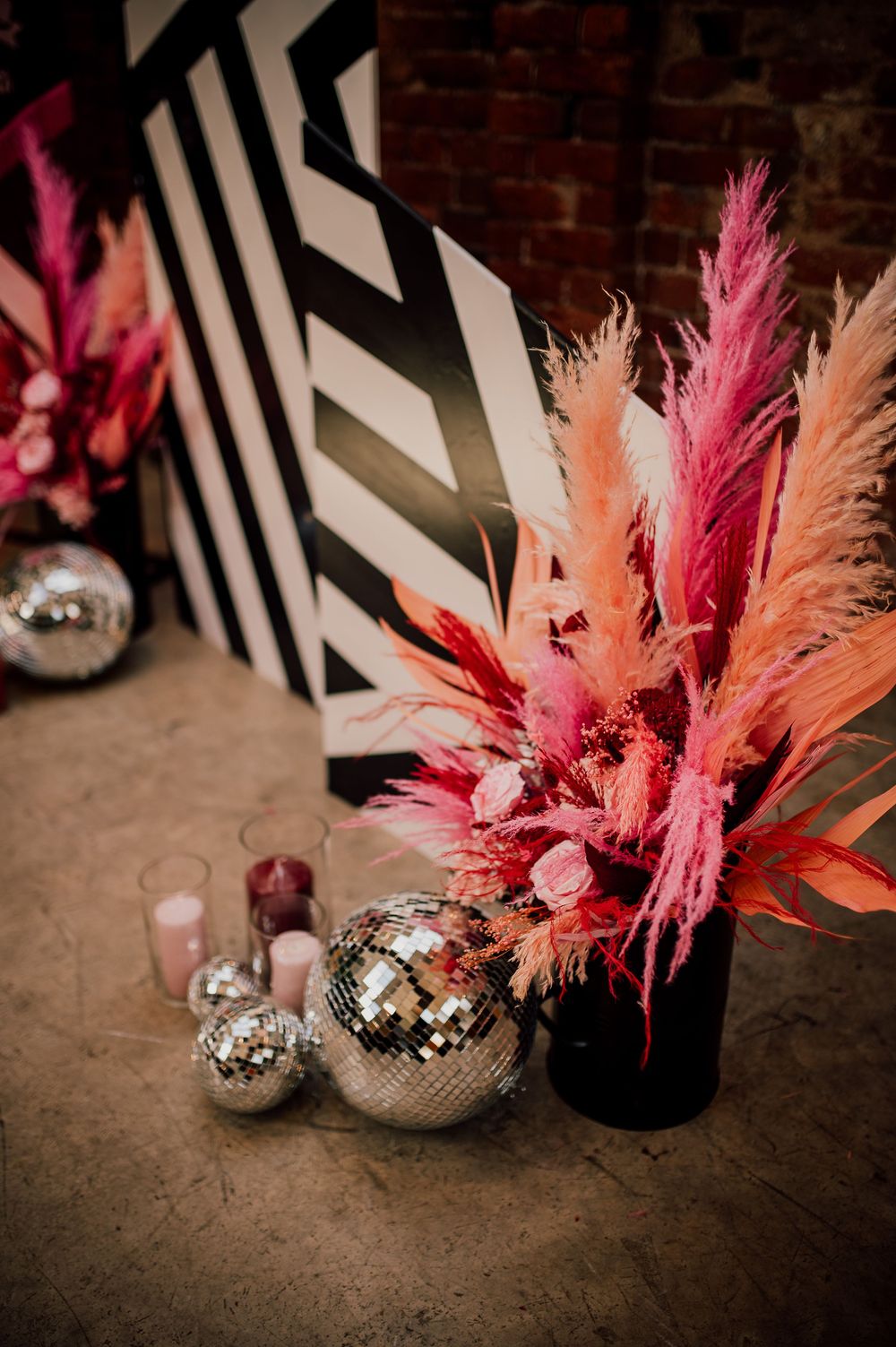 Disco ball ornaments and pink pampas grass decorations arranged against a dark brick wall create a modern party atmosphere.