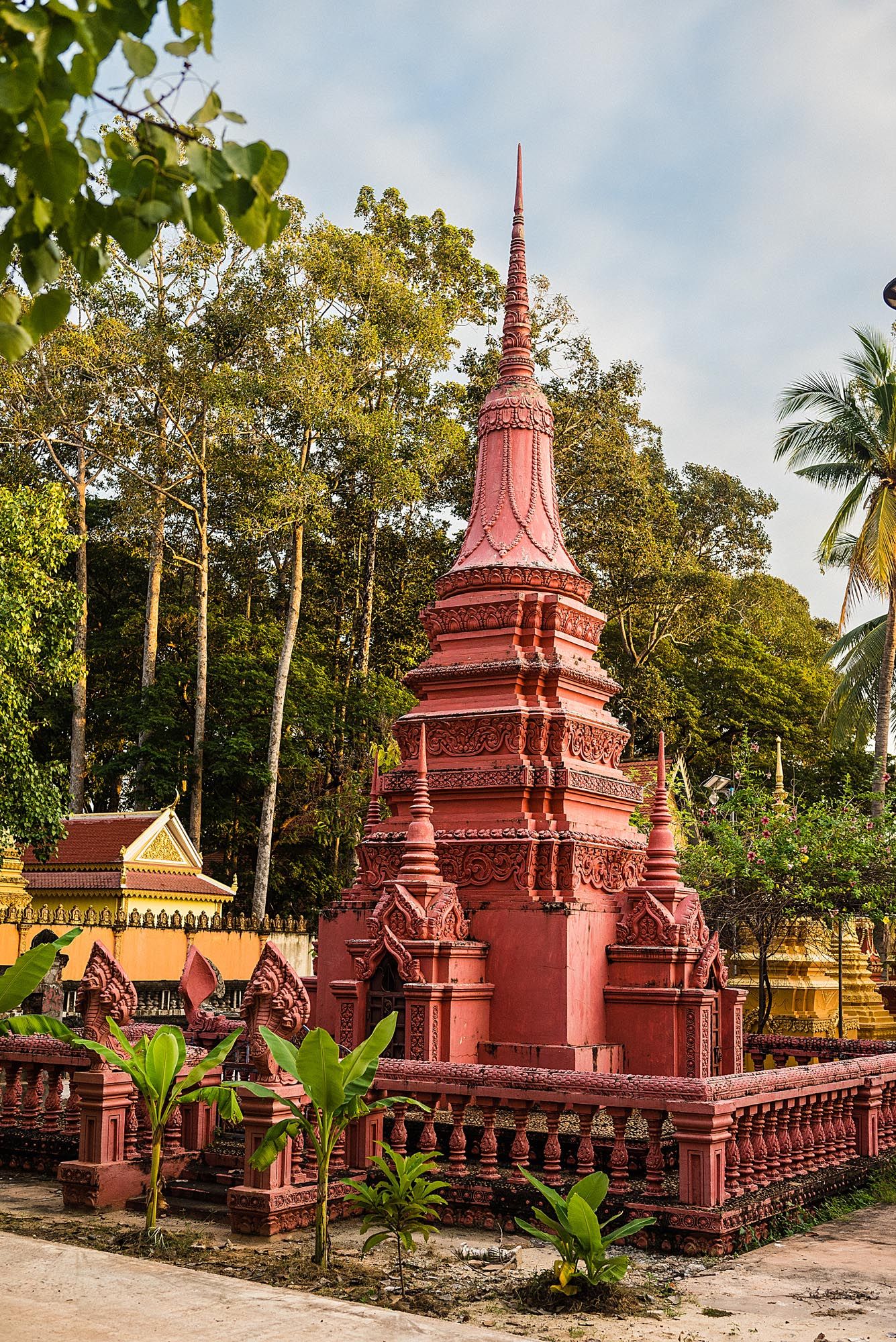 Red Buddhist temple stupas with ornate architectural details stand among lush tropical greenery and palm trees.