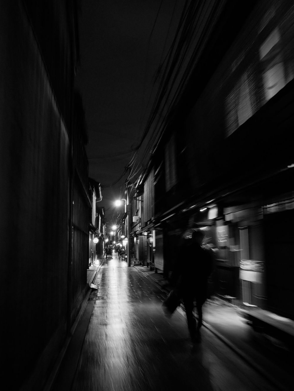 Moody black and white photograph of a narrow alley in Japan at night with glowing street lights.