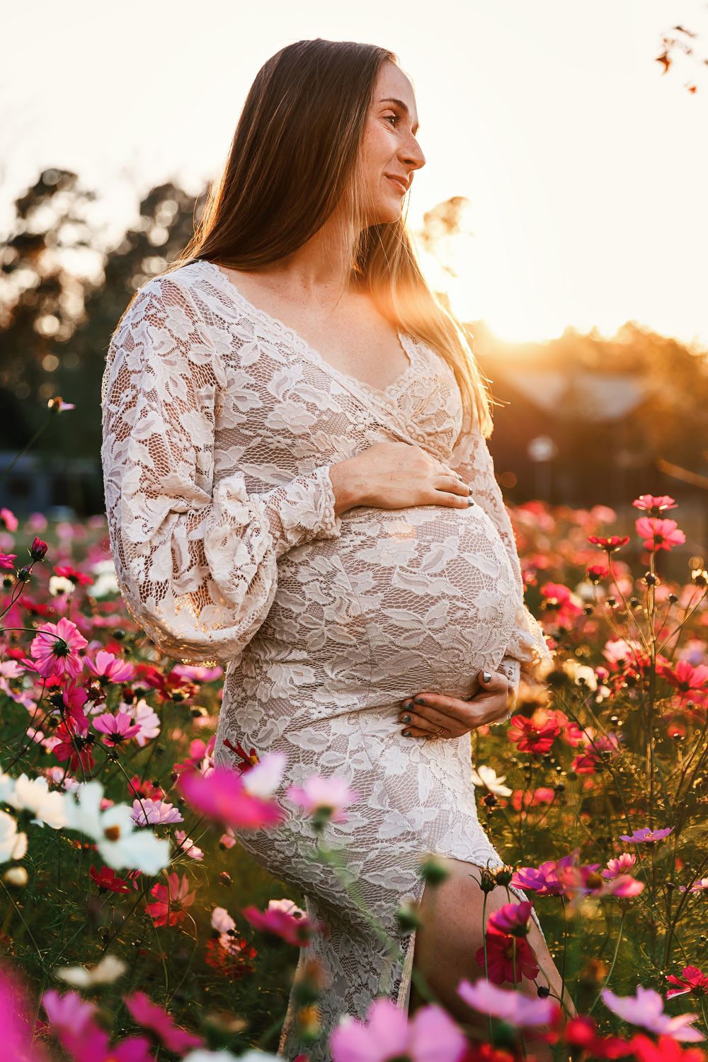 Expectant person in white lace dress poses in a field of pink cosmos flowers at golden sunset.