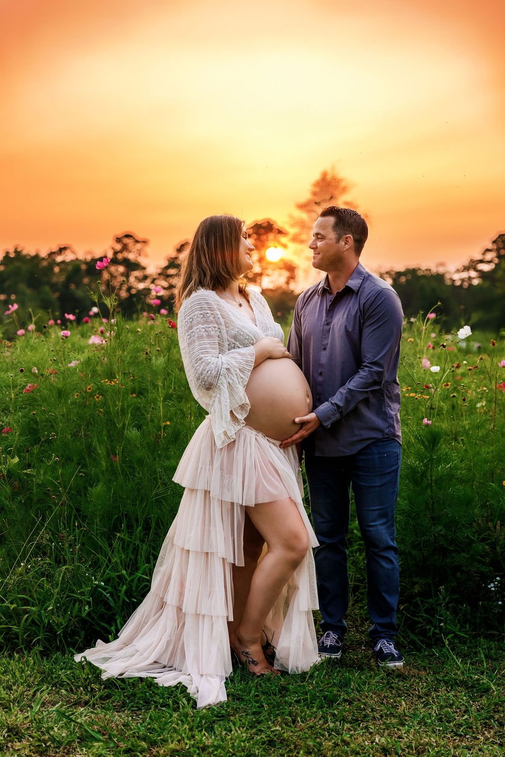A couple shares an intimate moment in a wildflower field at sunset, with the person wearing a flowing white lace dress.