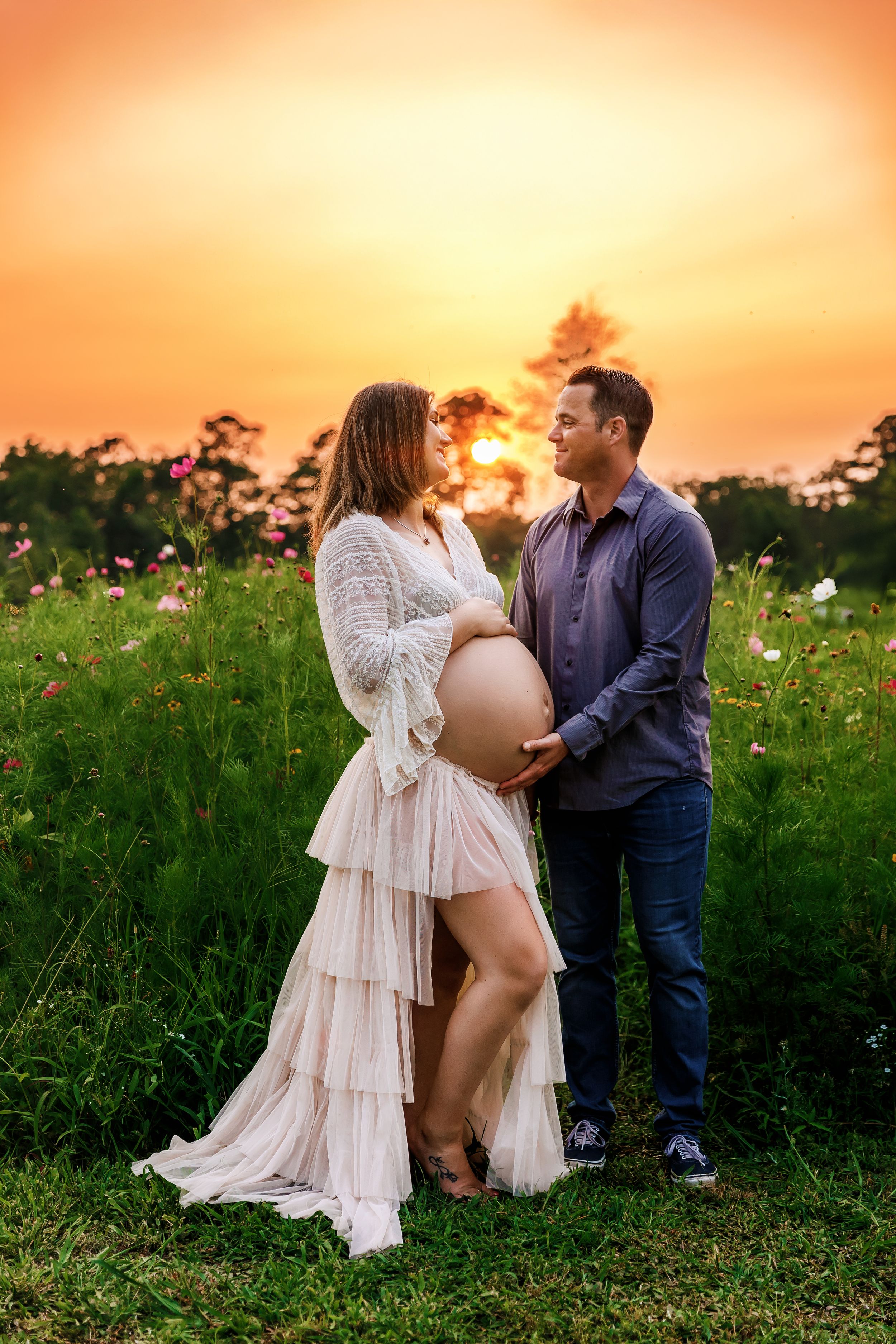 A couple shares an intimate moment in a wildflower field at sunset, with the person wearing a flowing white lace dress.