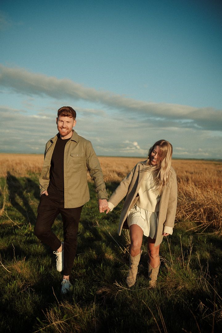 A couple holds hands while walking through a golden field at sunset under a turquoise sky.