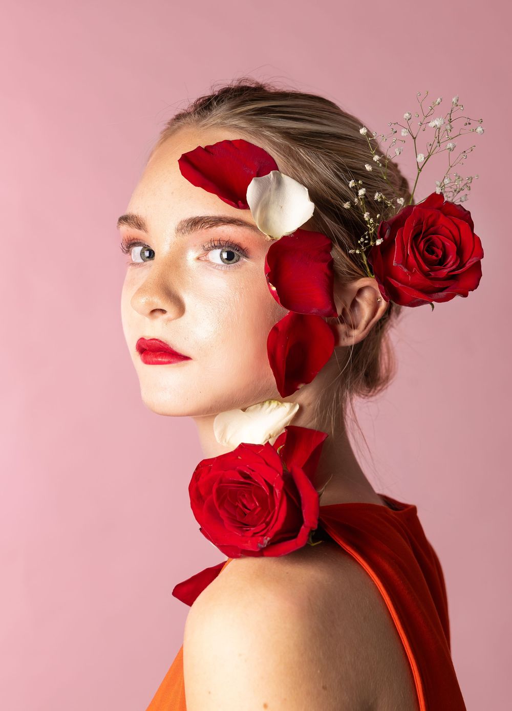 A girl in front of a pink background with rose petals on her face and head, looking straight