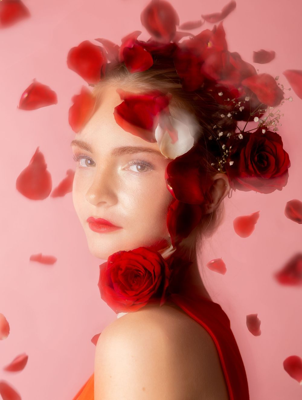 A girl in front of a pink background with rose petals falling around her, looking straight