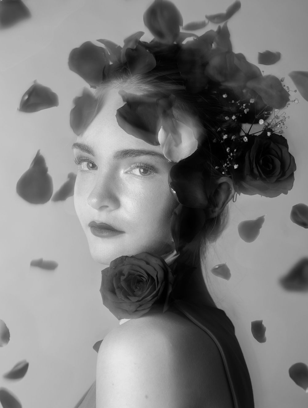 Black and white photo of a girl in front of a pink background with rose petals falling around her, looking straight