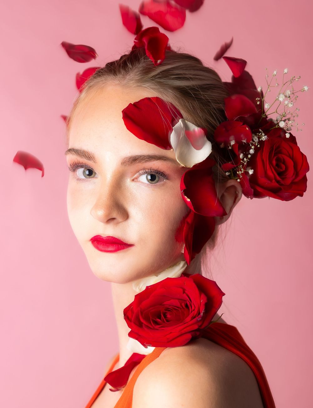 A girl in front of a pink background with rose petals slightly falling on her head, looking straight
