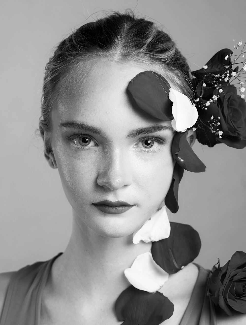 Black and white photo of girl in front of a pink background with rose petals on her face and head, looking straight with neutral expression