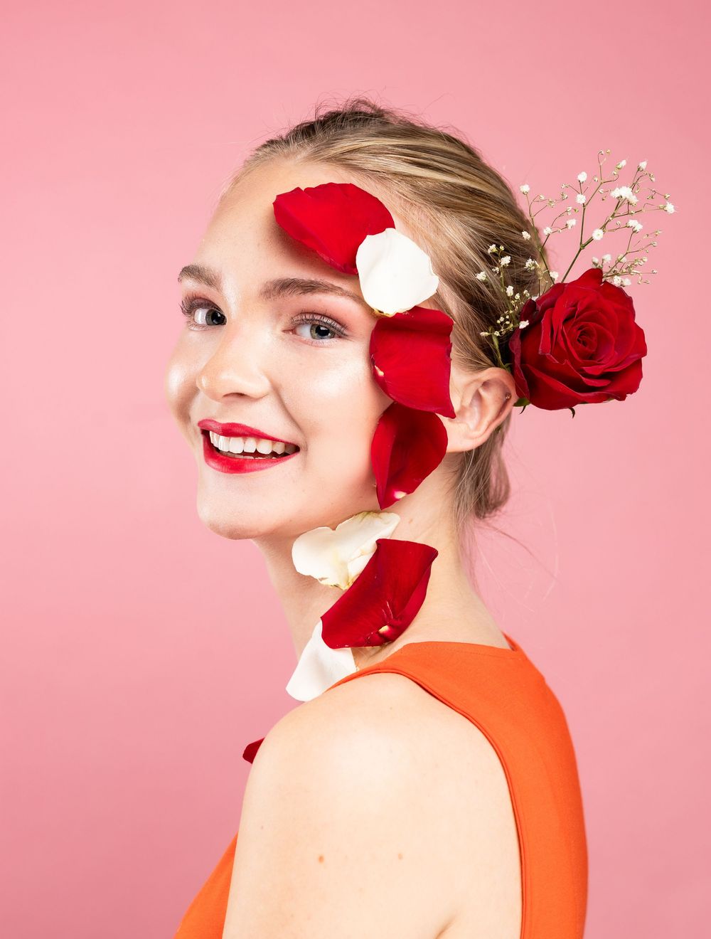A girl in front of a pink background with rose petals on her face and head, looking straight and smiling