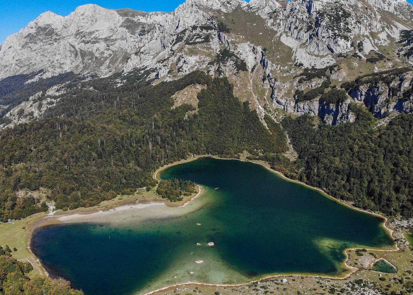 Aerial panoramic view of a turquoise alpine lake surrounded by rugged mountain peaks and forested slopes in summer.