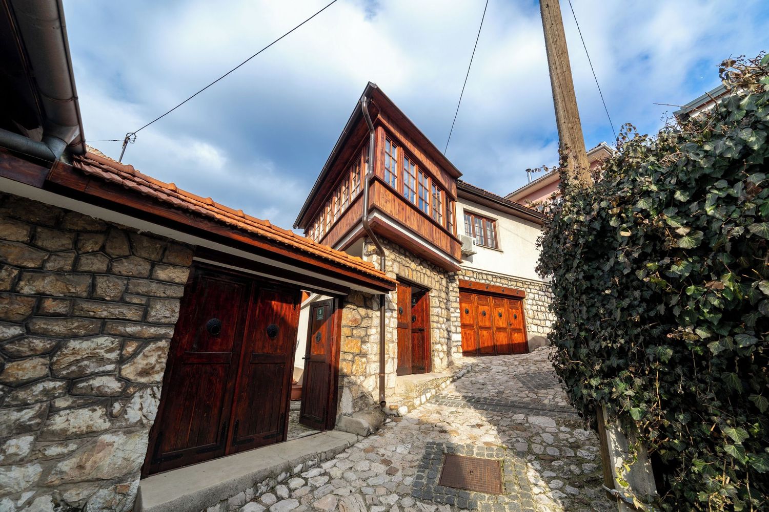 A narrow cobblestone street winds between traditional stone houses and red-tiled roofs in a Mediterranean village.