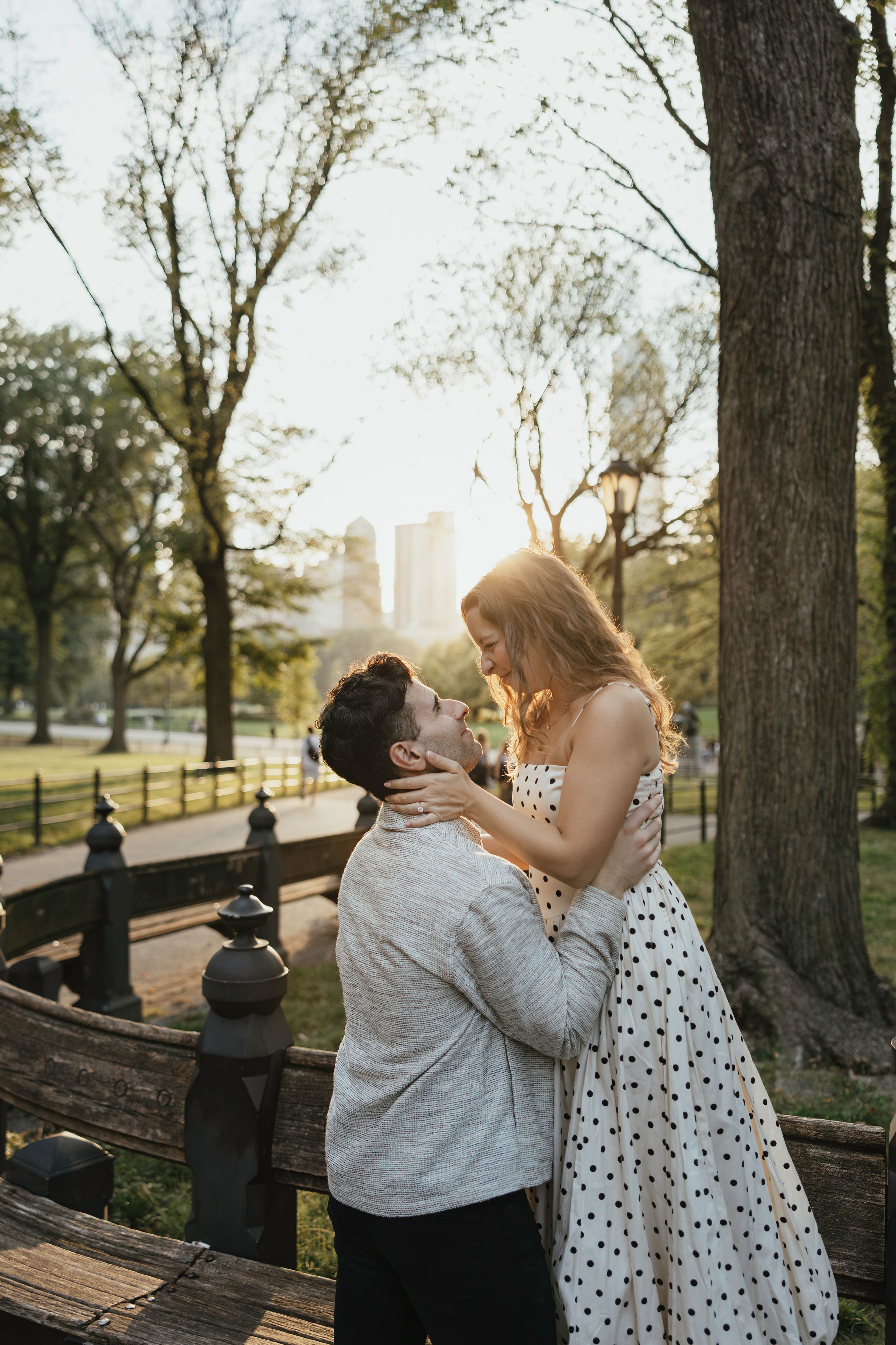 A romantic couple shares an intimate moment on a park bench at sunset with city buildings visible in the background.