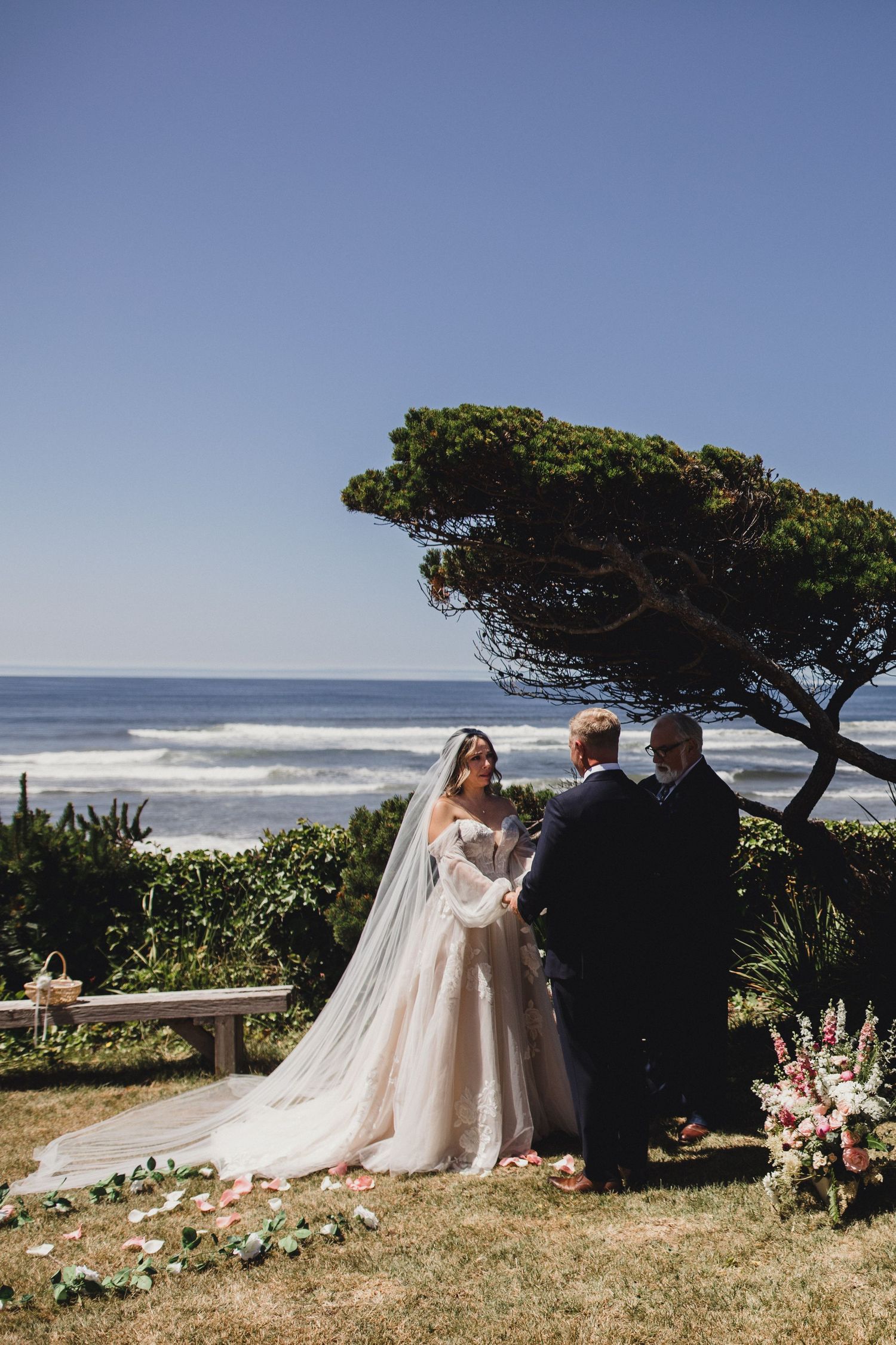 A romantic coastal wedding ceremony overlooking the Pacific Ocean with windswept cypress trees and floral decorations.