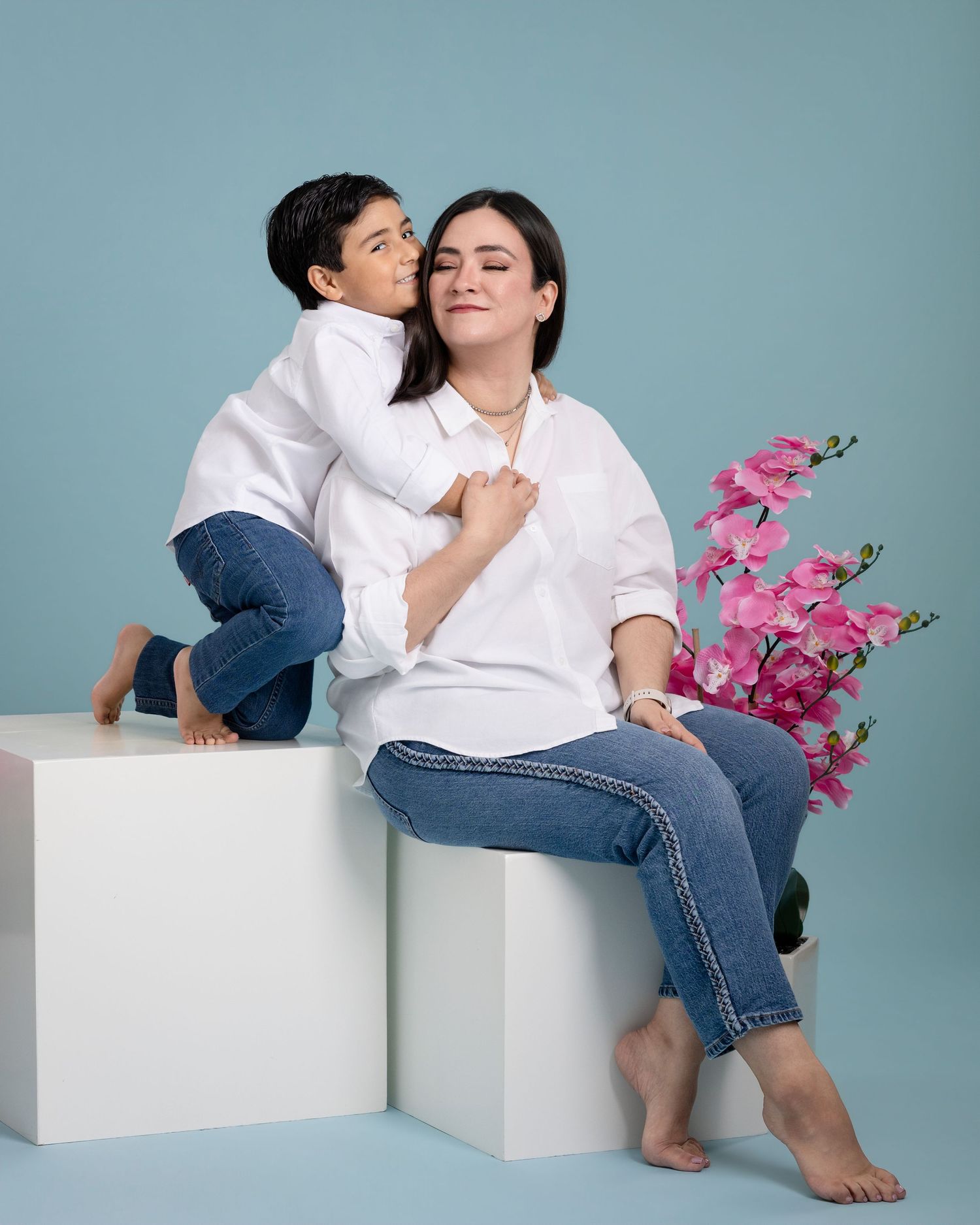 Mother and child in white shirts and jeans pose playfully on white blocks against blue backdrop with pink orchids.