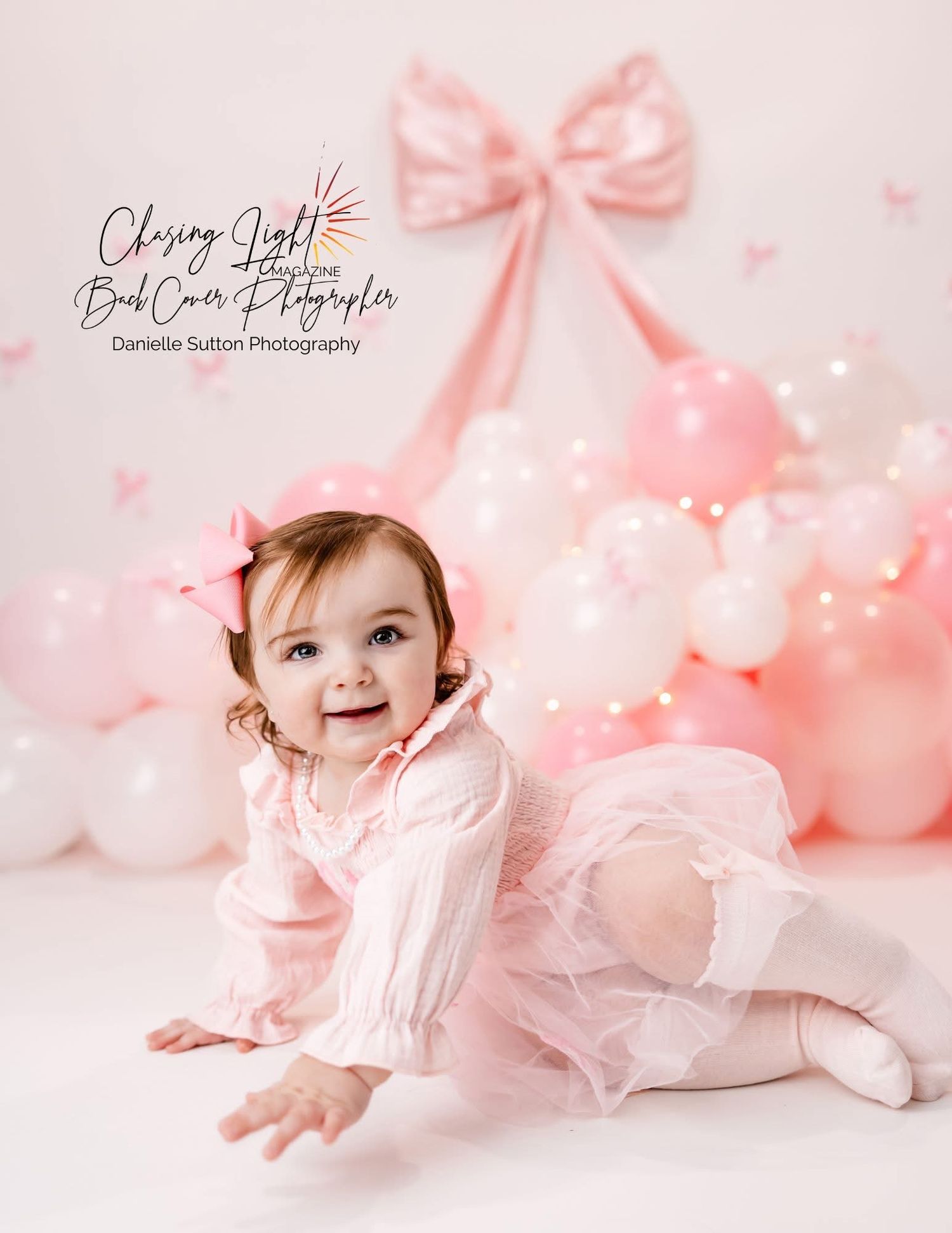 Adorable baby girl in pink tutu sits among pink balloons and bow backdrop in professional portrait.