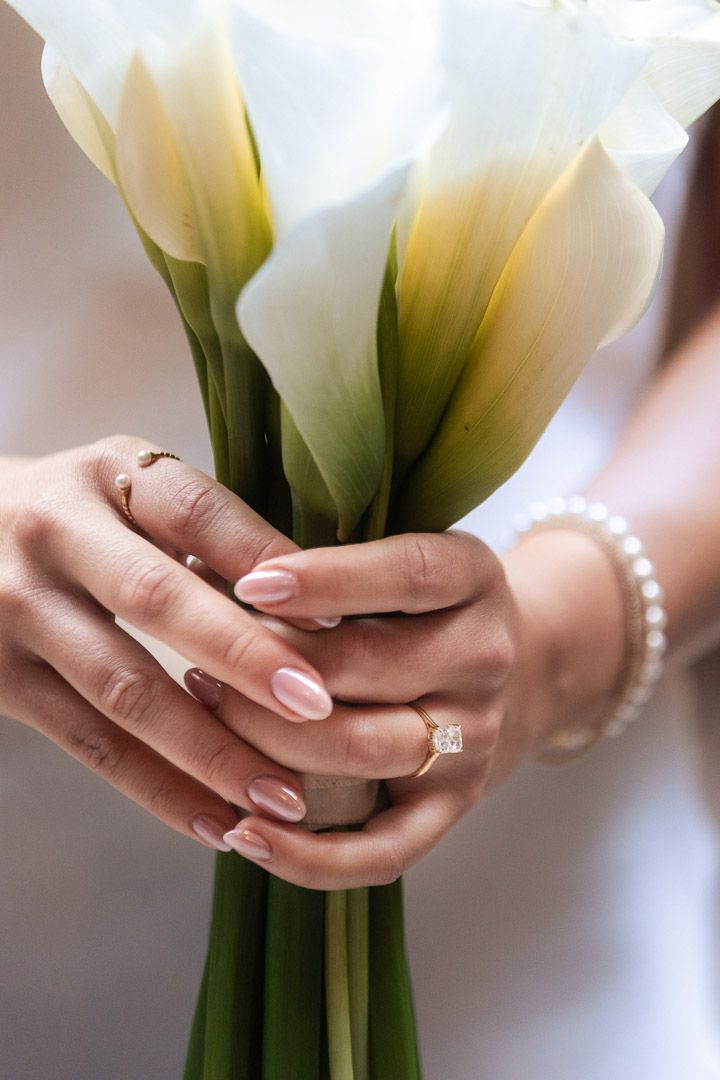A bride's hands gently cradling delicate white calla lily flowers against a soft blurred background.