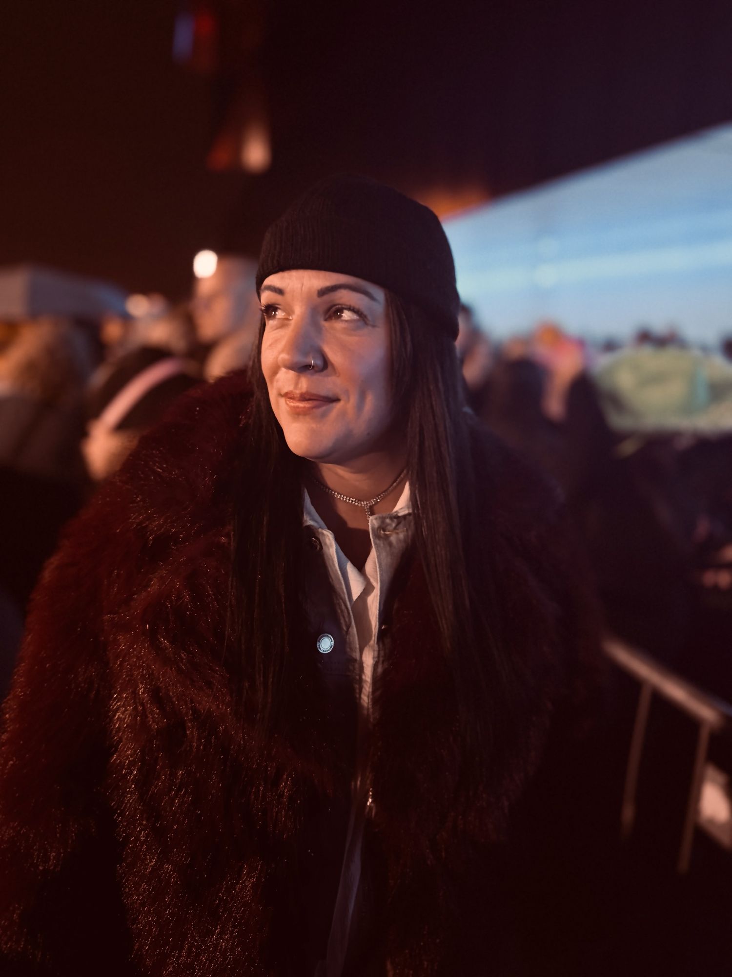 Person wearing a dark beanie and burgundy fur coat looking to the side at an evening event.