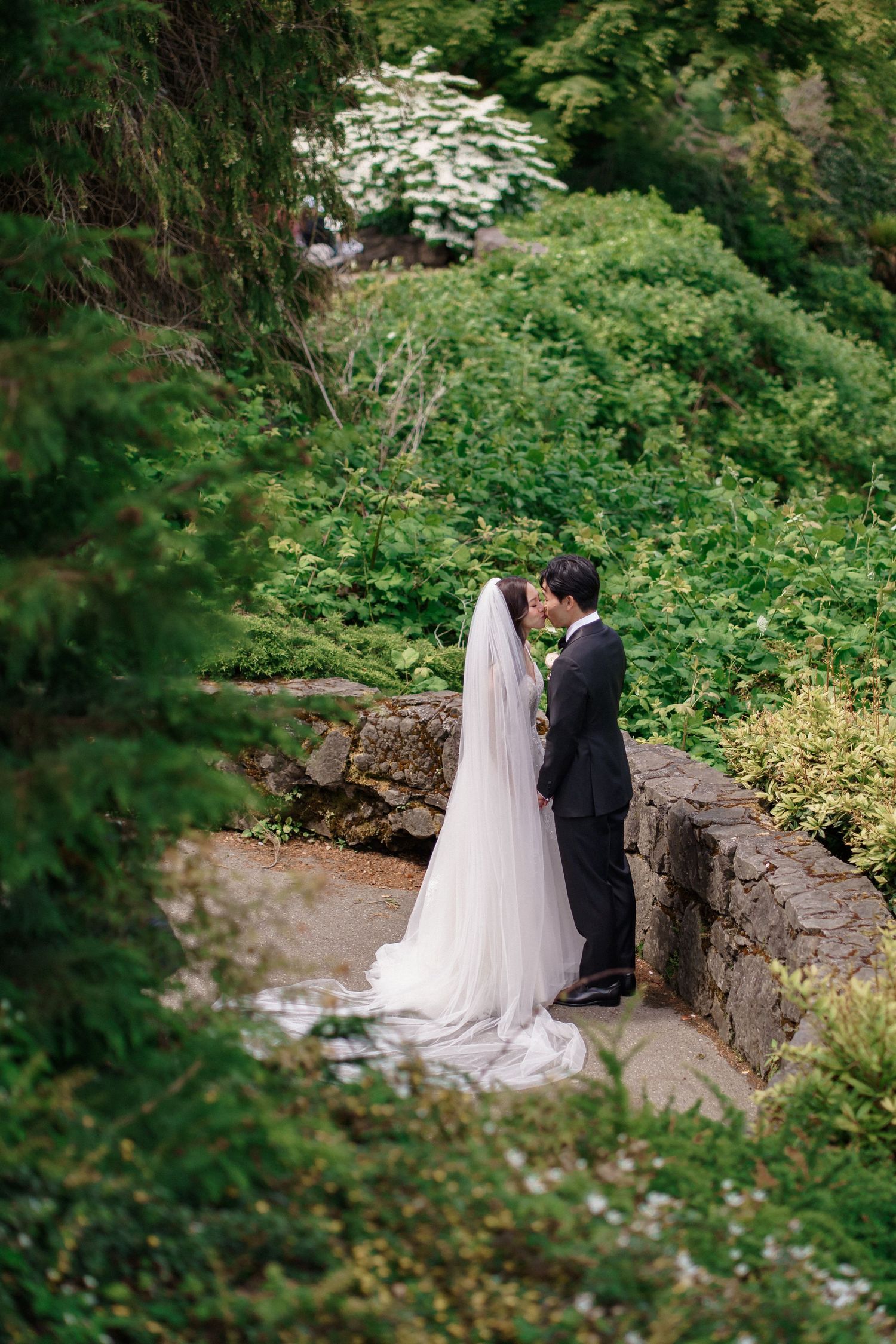 A couple in wedding attire stands together in a lush green garden setting with stone walls and natural foliage, Queen Elizabeth Park Wedding