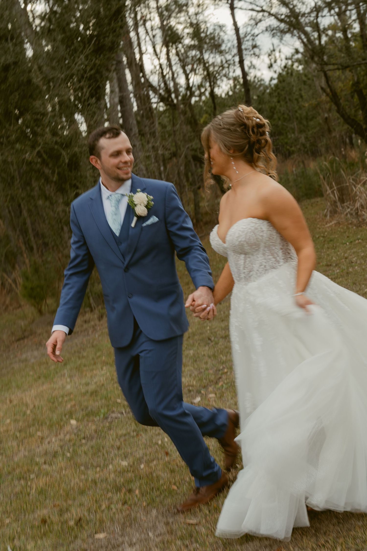 A couple in wedding attire holding hands and walking together through a wooded outdoor setting.