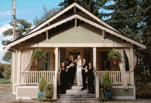 A rustic white chapel with a covered porch and cross on top stands surrounded by trees and greenery.