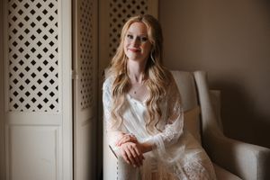 A woman in a white lace dress poses near a window with lattice details in a romantic indoor setting.
