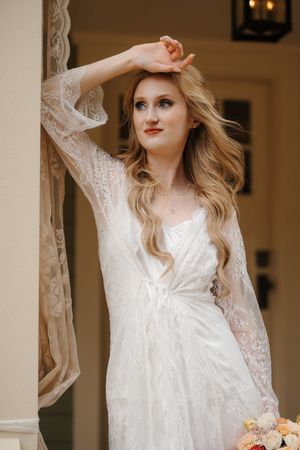 A bride in a delicate white lace robe poses elegantly against a light-colored pillar holding a peach floral bouquet.