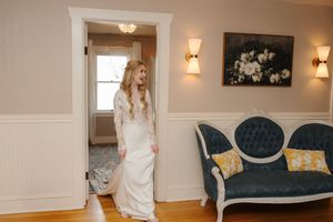 A bride in a white wedding gown stands in an elegant room with white walls and wall sconces.