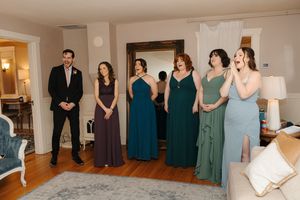 A wedding party wearing coordinated blue and teal dresses stands in line against a light colored wall during a celebration.