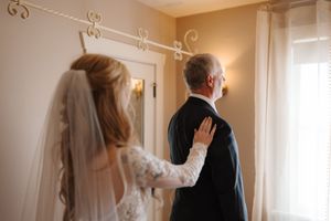 A bride in a lace gown and veil touches her father's shoulder during a first look moment before the wedding ceremony.
