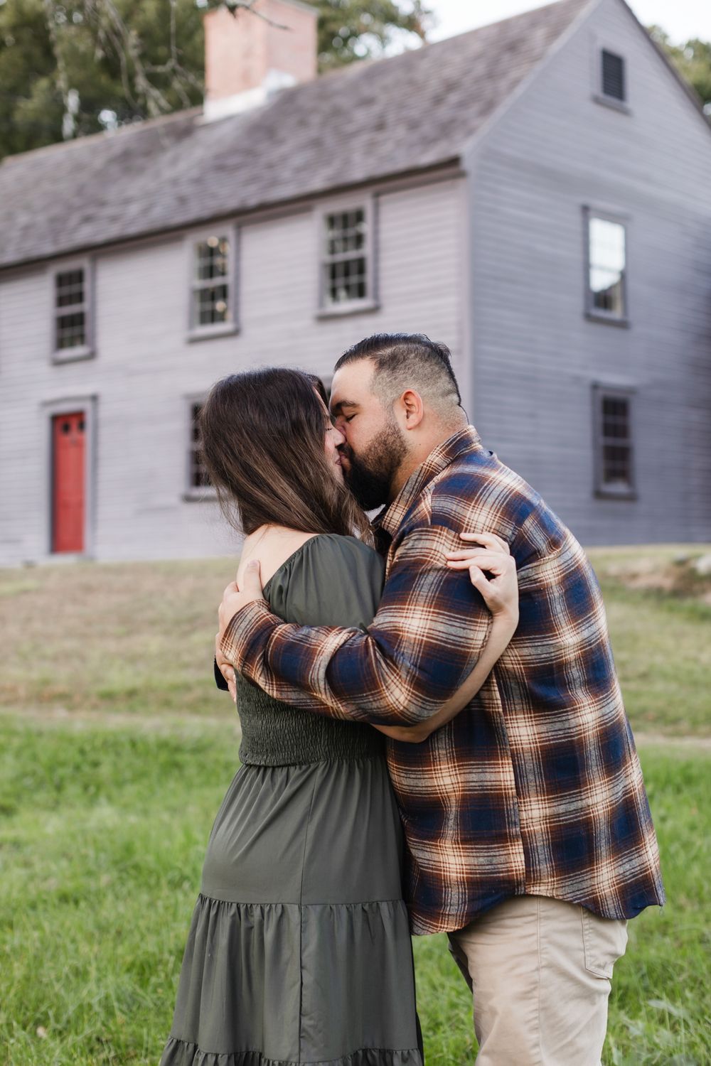 A man in plaid flannel kisses his fiancee wearing a green dress in front of Meriam House in Concord, MA for engagement photos.
