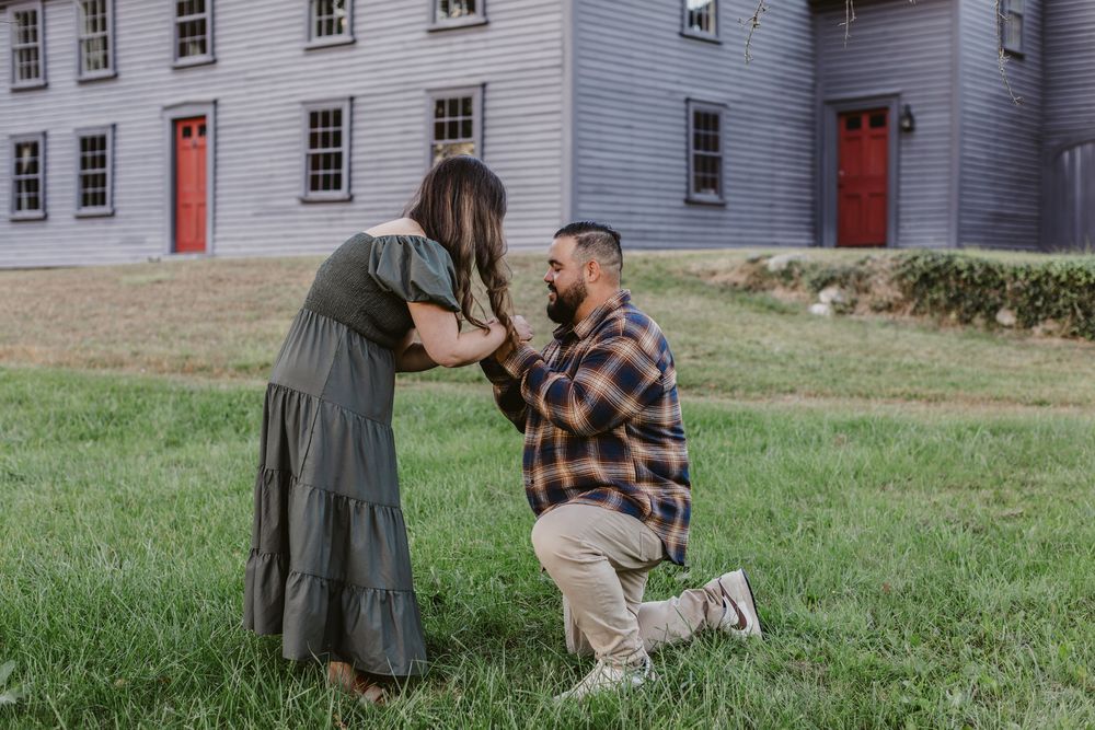 Man wearing flannel shirt proposes and bends knee to woman in green dress in front of Meriam House in Concord, MA for engagement photos.