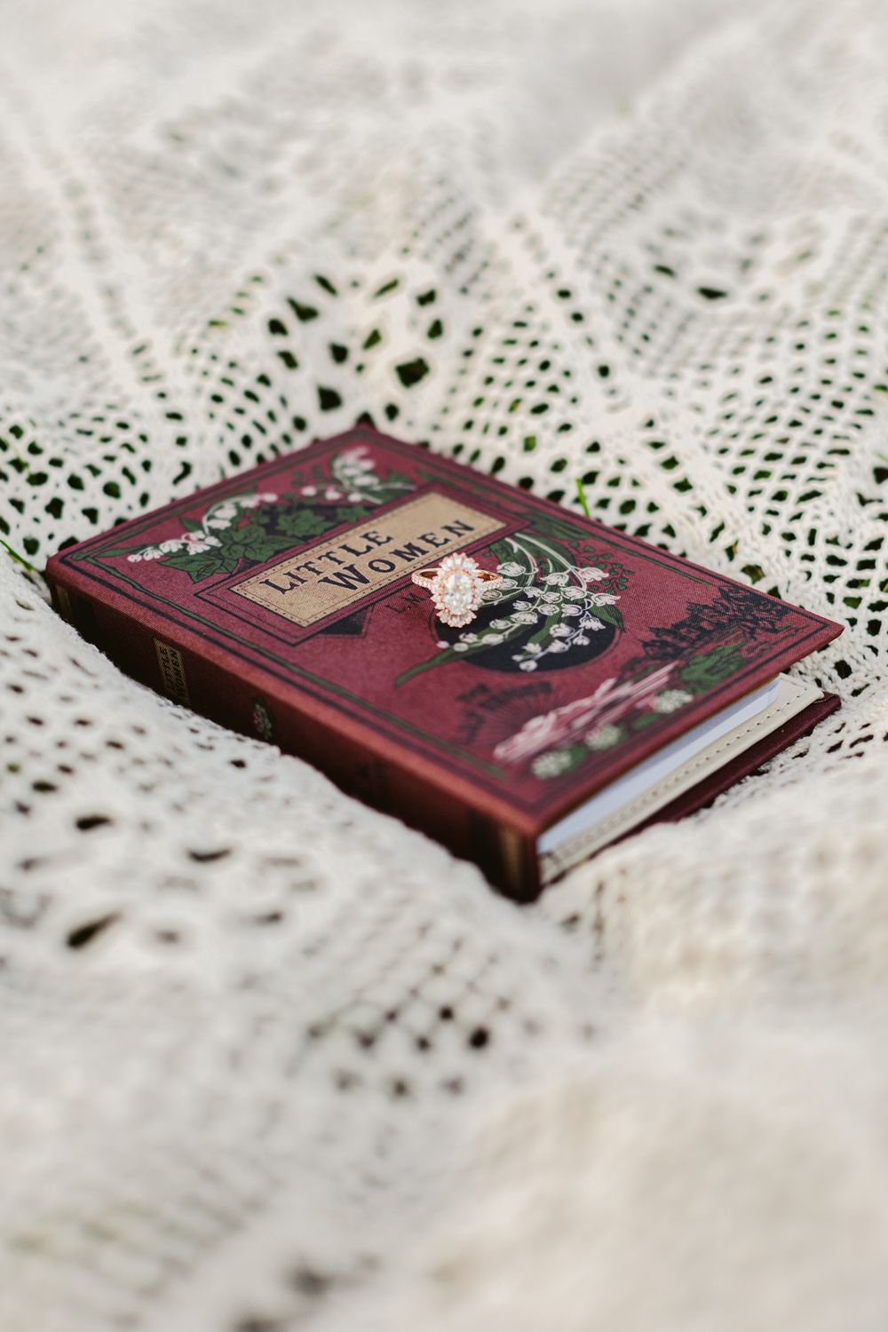 Little Women book wallet lays on a cream colored crochet blanket with a vintage gold engagement ring on it for engagement photos in Concord.