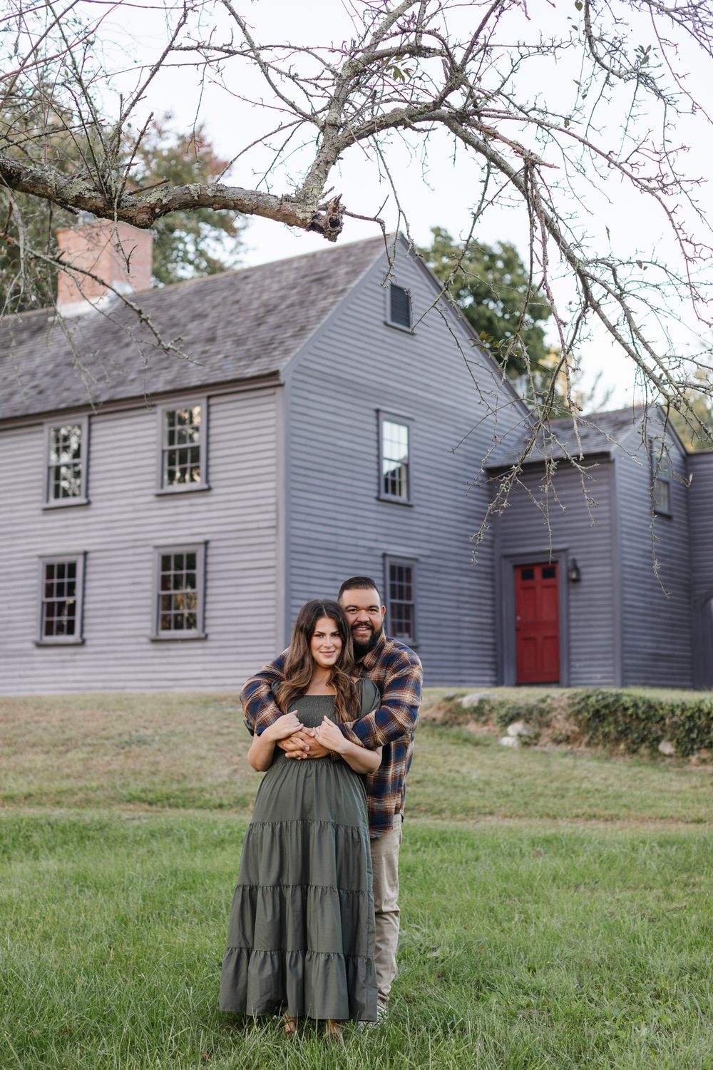 A man embraces his fiancee from behind in front of Meriam House in Concord, MA for engagement photos. Southern Maine engagement photographer