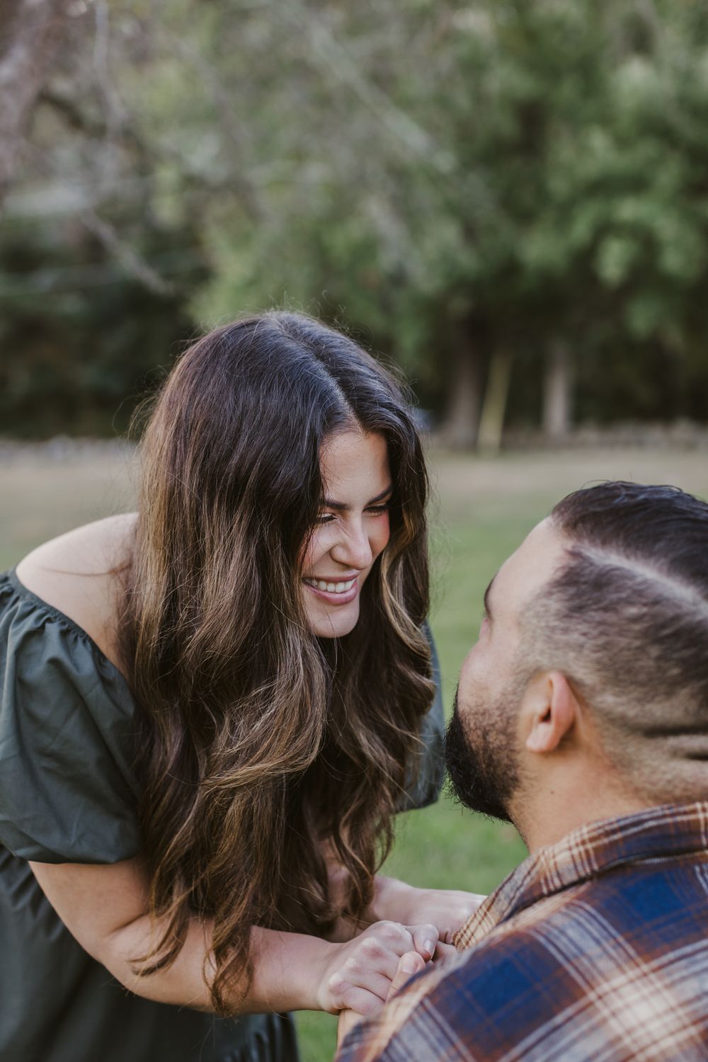 A woman with long brown hair and green dress smiles joyfully at man who proposed to her wearing plaid flannel in Concord, Massachusetts.