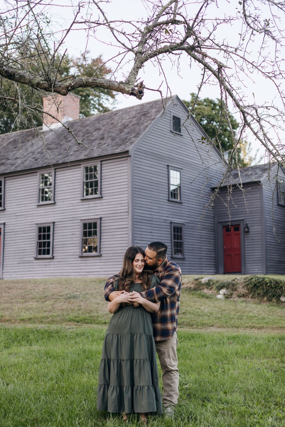 A man kisses his fiancee's temple in front of Meriam House in Concord, MA. North Conway, Woodstock, White Mountains engagement photographer
