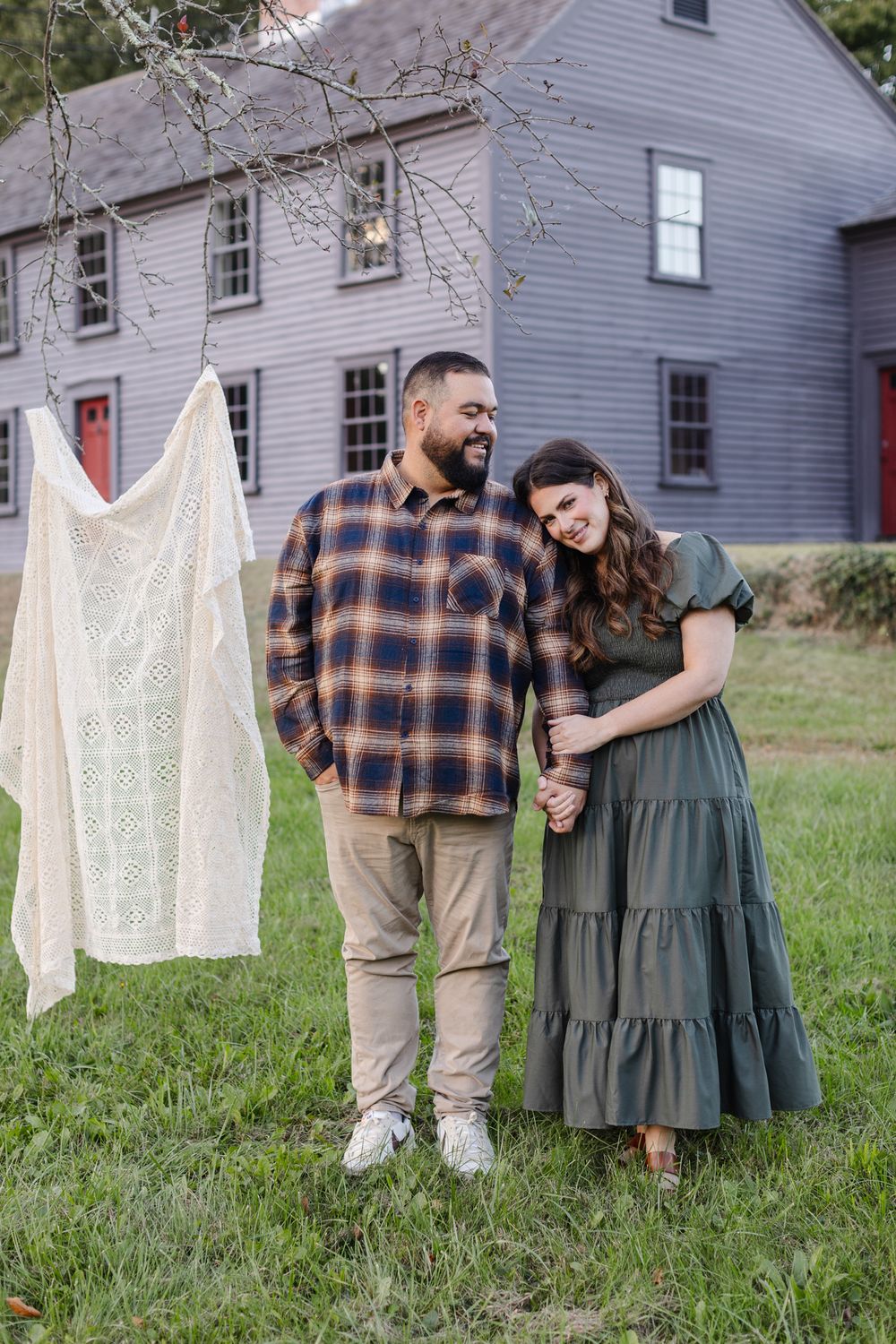 Woman in green dress rests head on shoulder of man wearing flannel shirt standing in front of historical house in Concord for engagement pic
