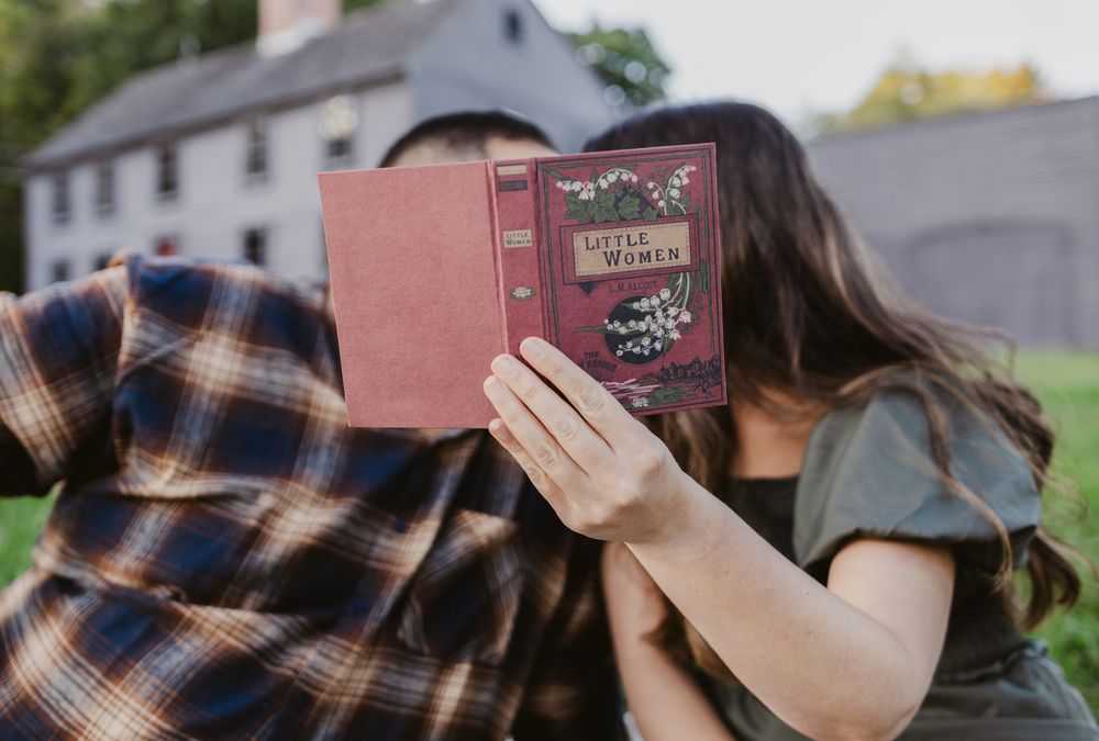 A woman in green dress holds up a Little Women book in front of fiancee's and her face in front of Meriam House in Concord for couple photos