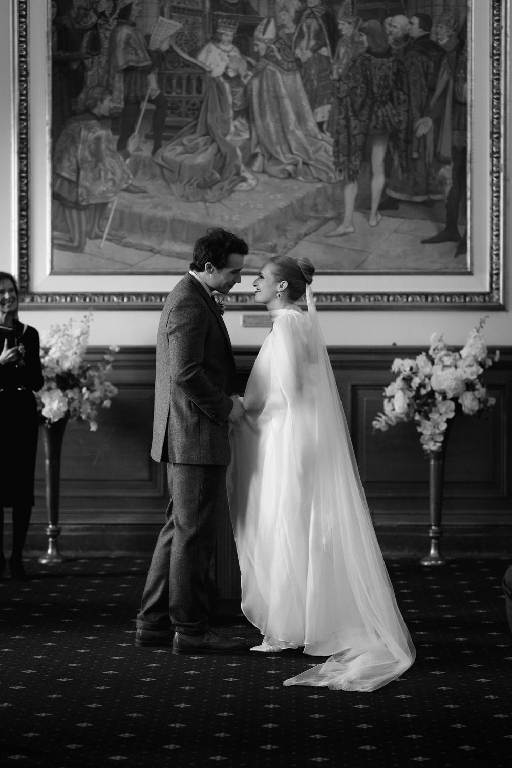 A couple shares an intimate dance moment in a grand hall with historic murals and elegant decor in black and white.