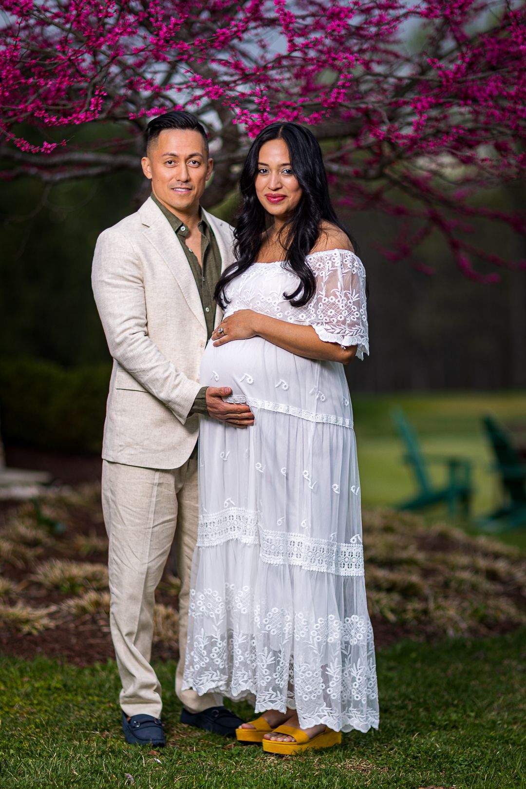 A person in a white lace maternity dress poses in front of blooming pink cherry blossoms in a garden setting.