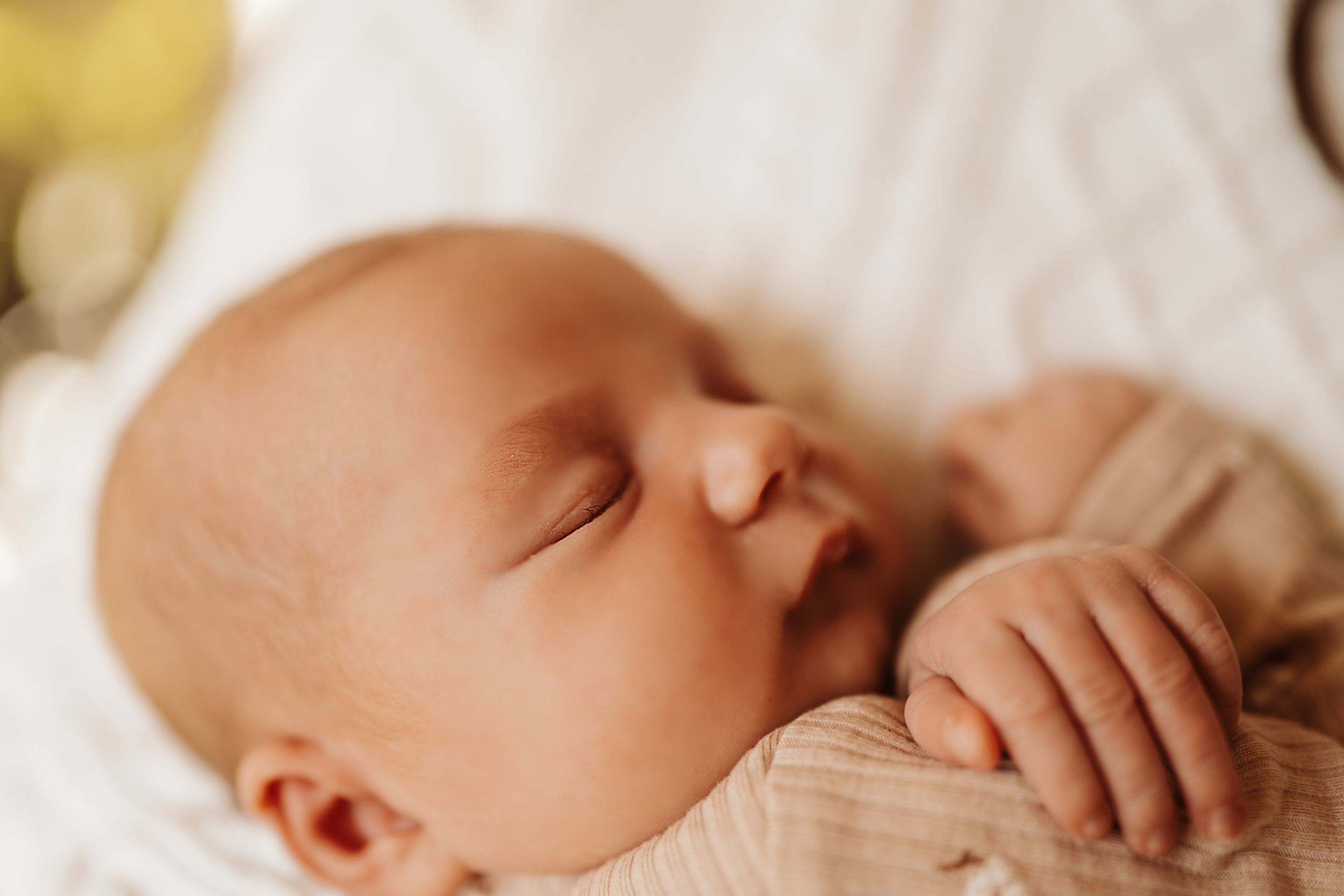 A sleeping baby rests peacefully on soft white bedding while being gently touched by caring hands.