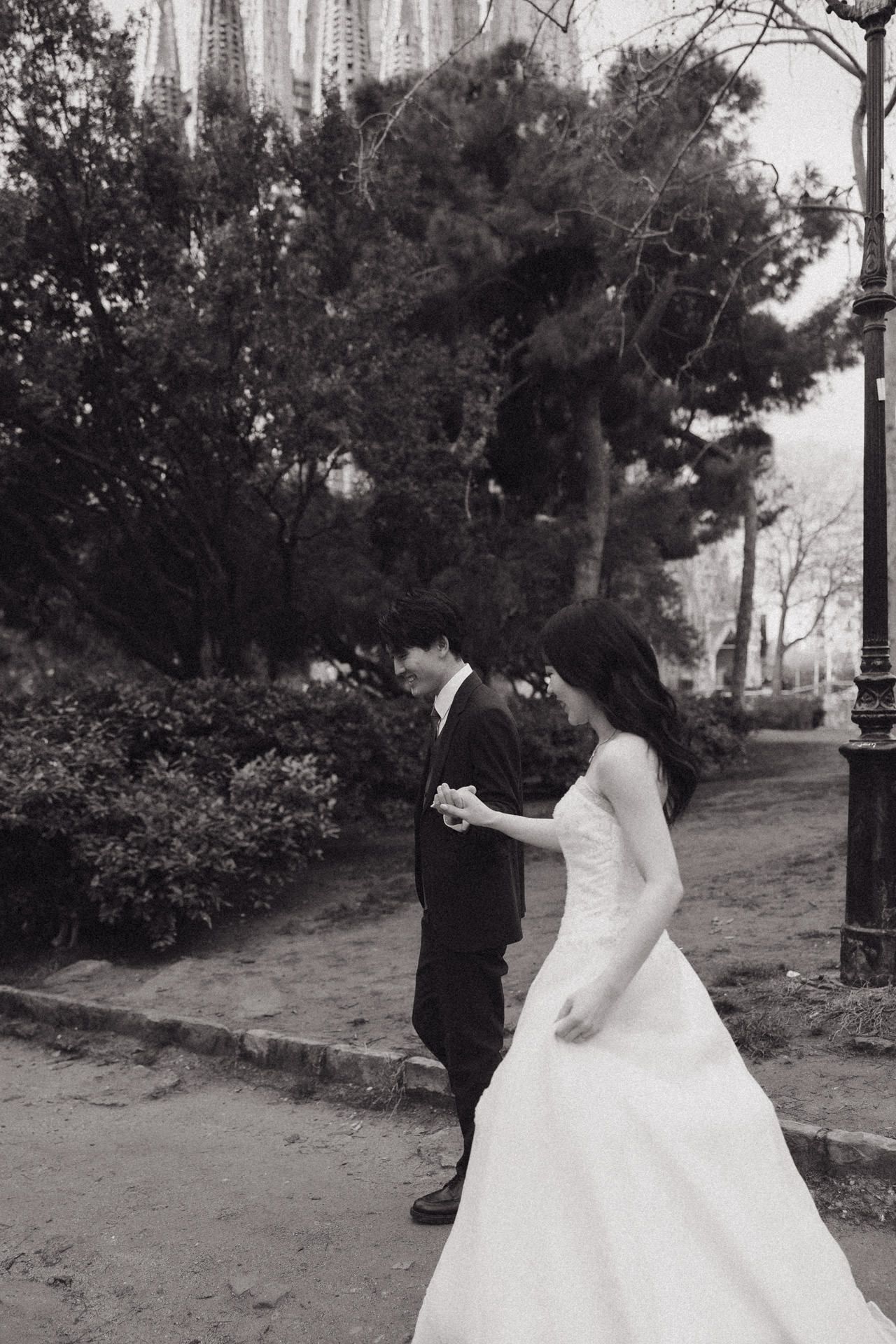 A couple in wedding attire walks along a wooded path on their special day. Couple photoshoot in Barcelona Sagrada Familia at sunrise