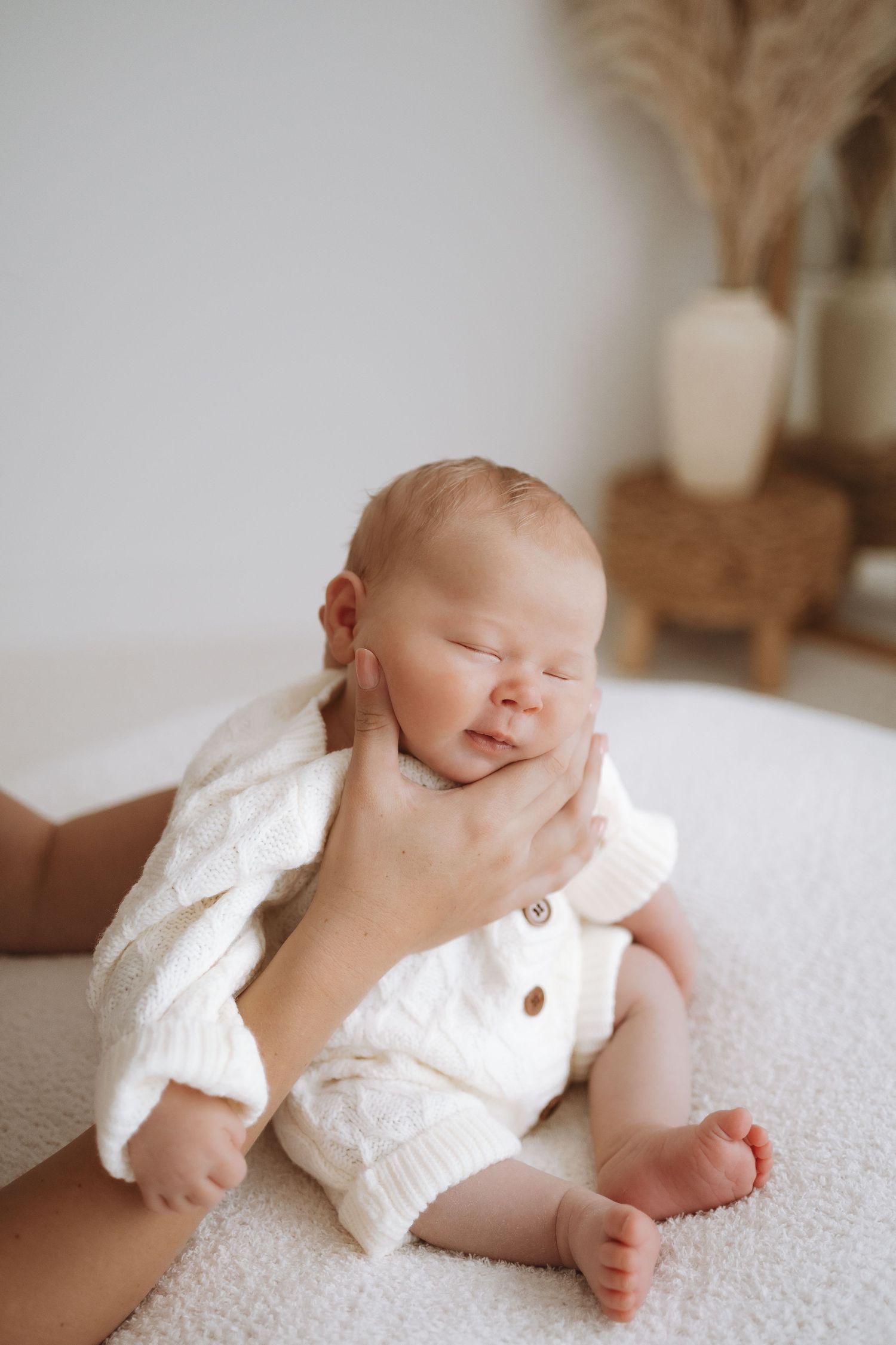 A small baby in white clothing sits on a light carpet while being supported by adult hands.