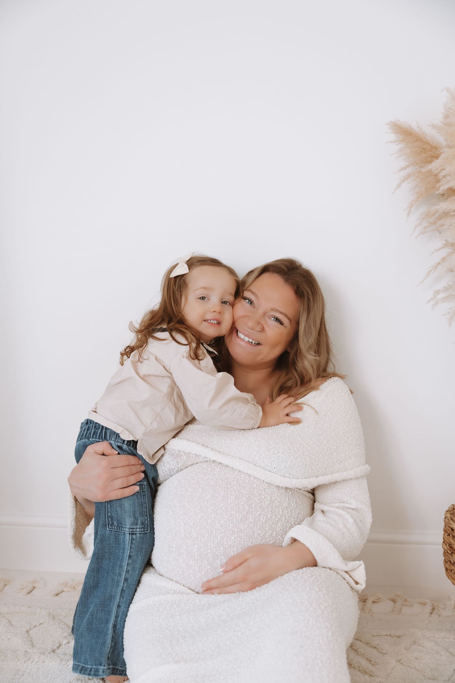A pregnant woman in a white sweater and jeans embraces a small child against a neutral background.