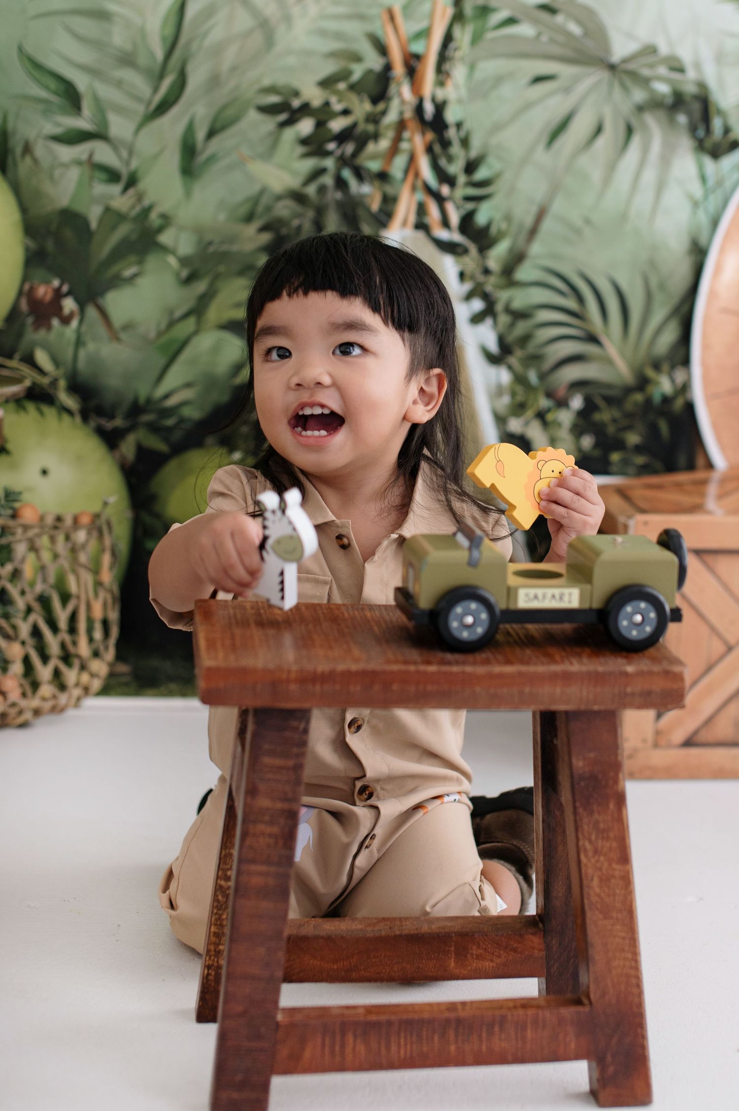 A small child in beige clothing sits at a wooden table with a tropical leaf backdrop.
