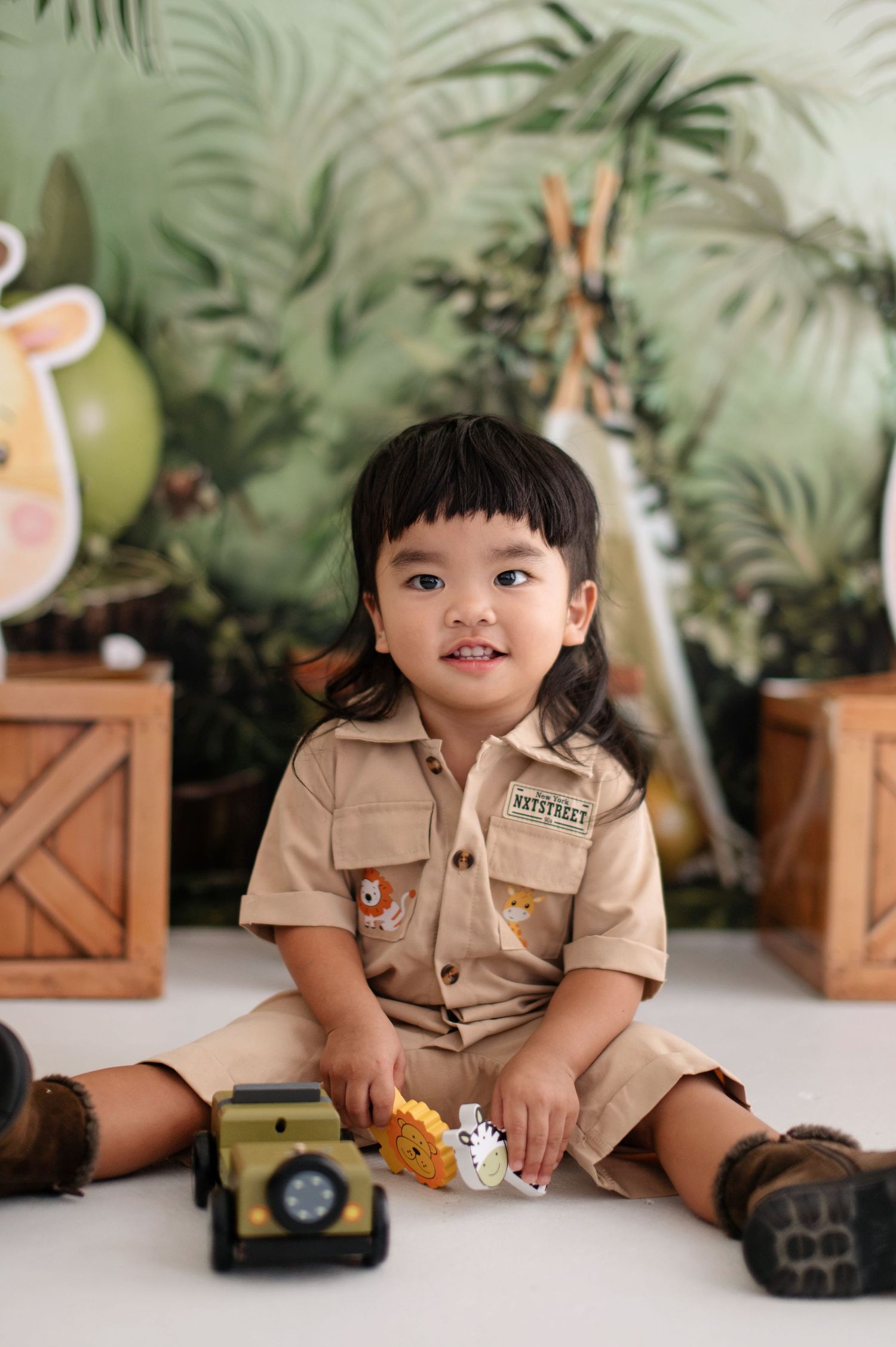 Child in safari ranger uniform plays with toy dinosaurs against tropical leaf backdrop in studio photoshoot series.