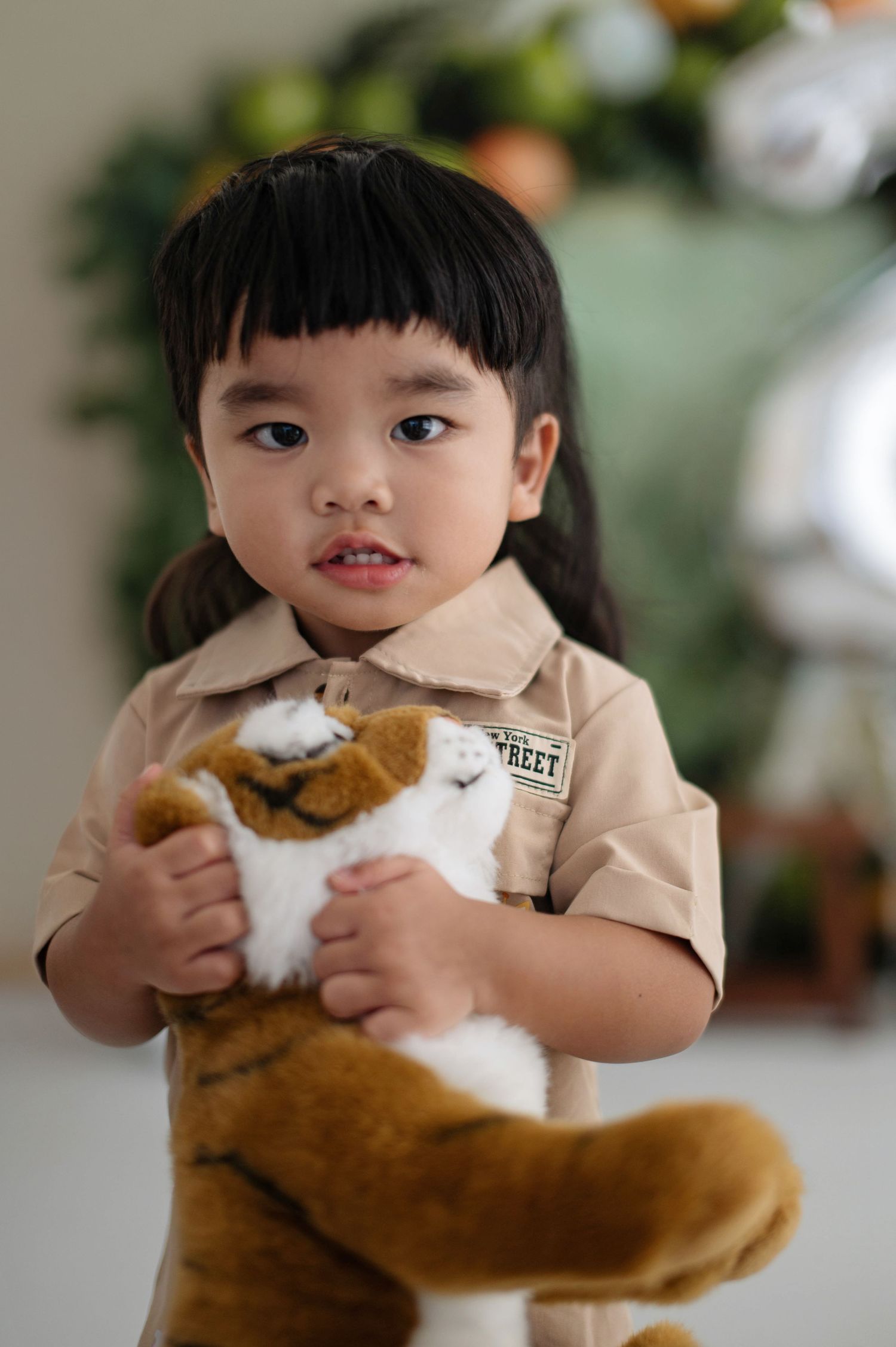 A small child in a beige uniform shirt holds a plush tiger toy against a blurred festive background.