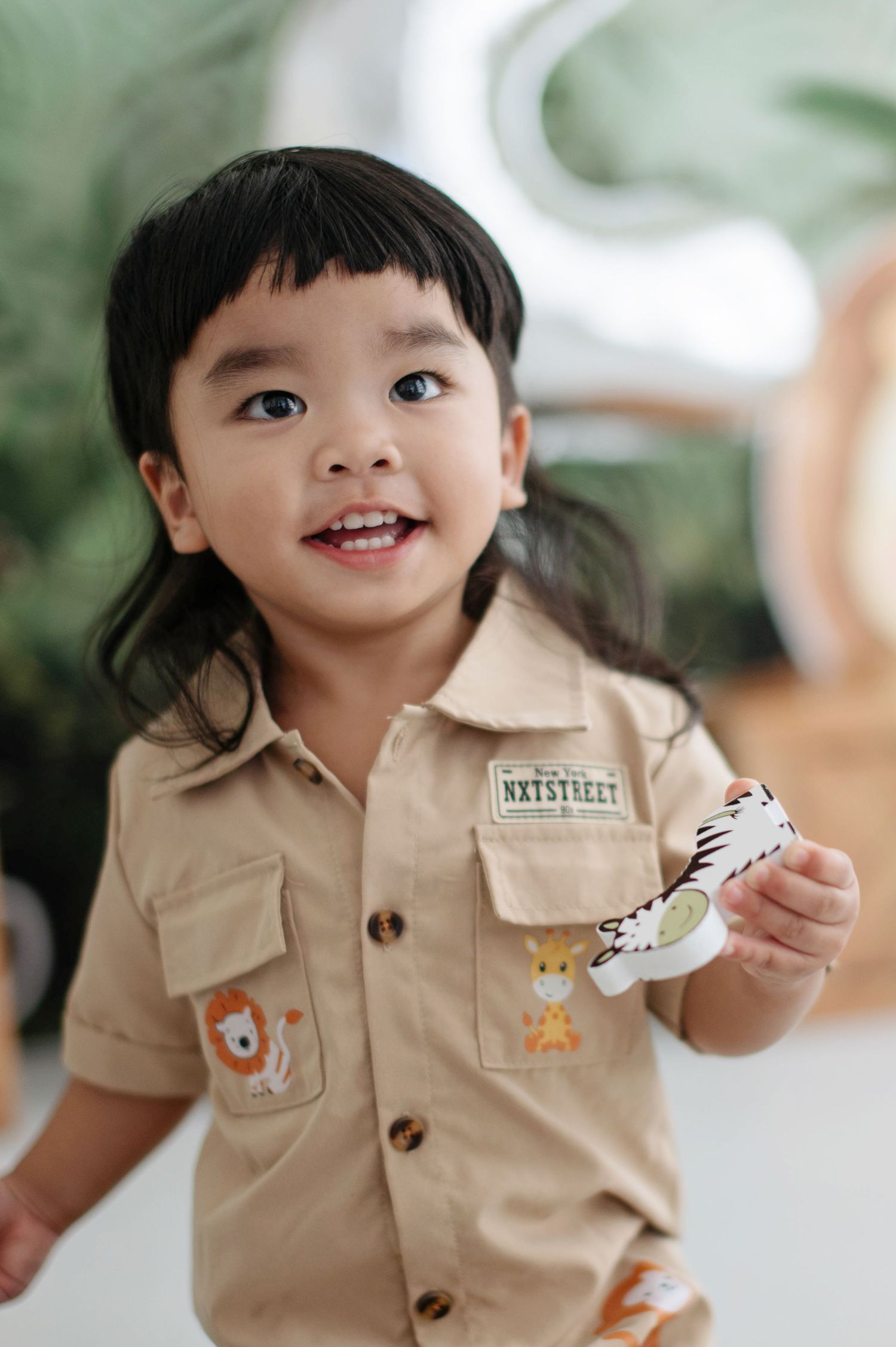Smiling child wearing a tan safari-style uniform with animal patches holds a toy while standing indoors.