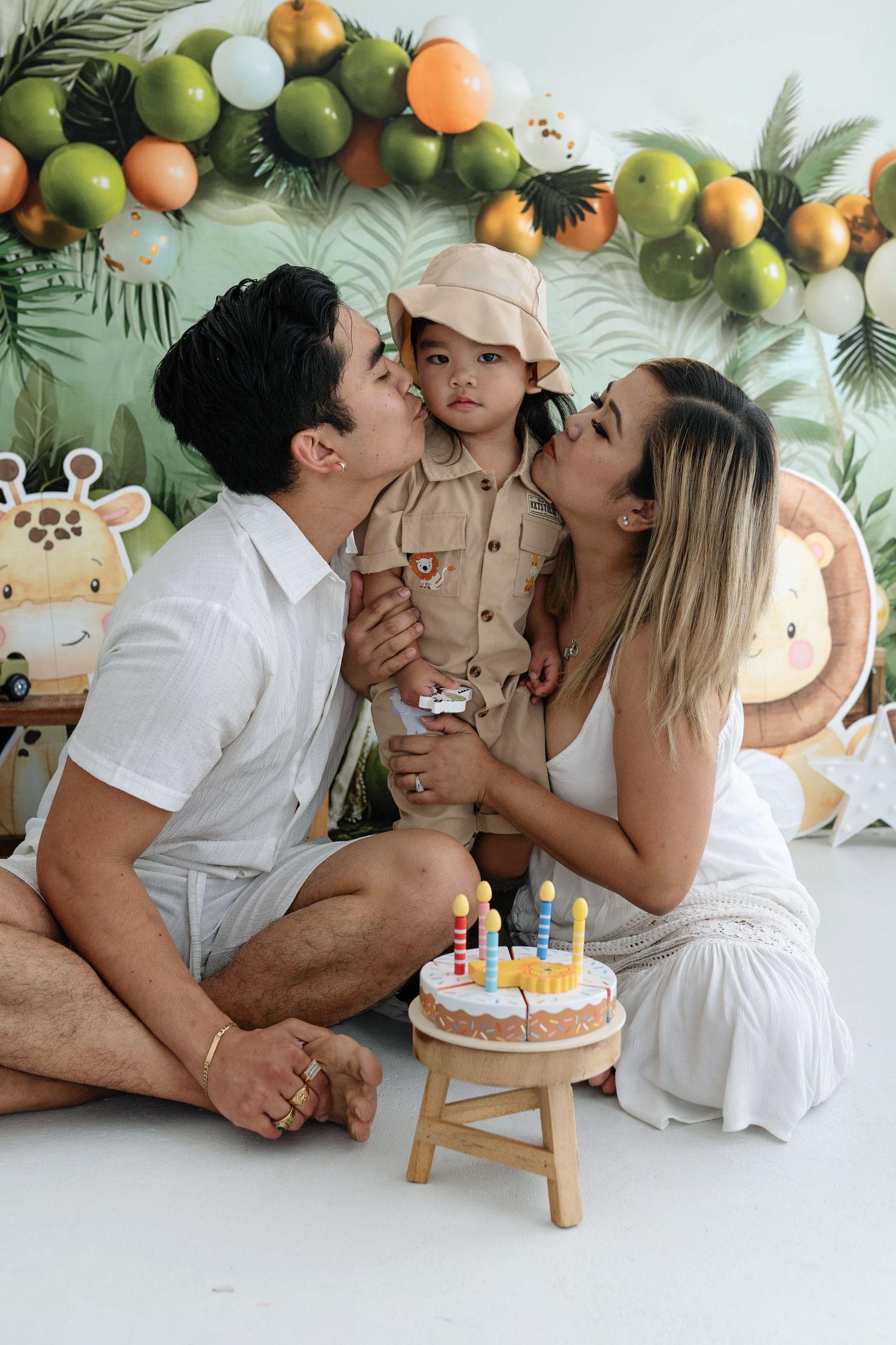 Family celebrates birthday with white cake and balloon decorations against tropical leaf backdrop with green and orange balloons.
